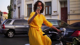 美丽微笑的女人穿着黄色长裙骑着一辆城市自行车，里面有一个篮子和鲜花，用她的手机拍摄。替身。Slowmotion视频素材模板下载