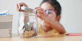 亚洲小女孩把硬币放进桌子上的一个透明的玻璃罐子里，比喻省钱的概念和声音选择集中在罐子上