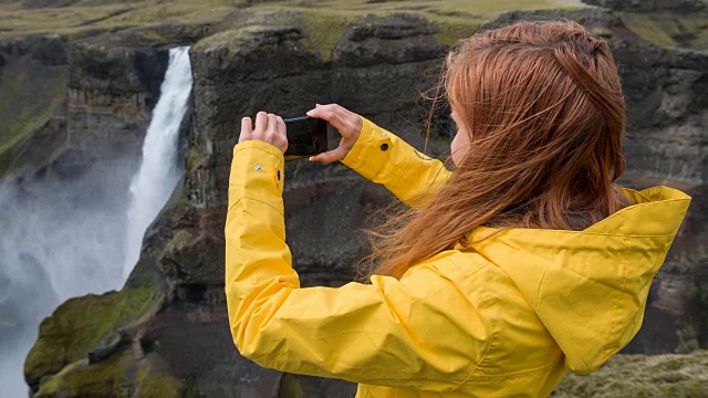 一名女子站在悬崖上，用智能手机拍摄瀑布