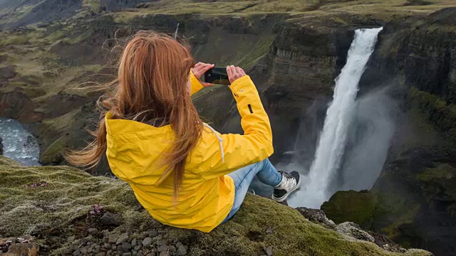 一名女子坐在峡谷边缘，拍摄瀑布的照片