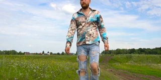 一个留着胡子，穿着破洞牛仔裤的男人在夏天沿着田野走着
