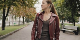 秋天来了。一个穿着酒红色皮夹克的女人走在城市的街道上，慢镜头