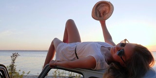 快乐的旅程，放松女孩在机器在海边，夏天的旅行，年轻女子躺在车顶在海边的河流，女性躺在汽车上