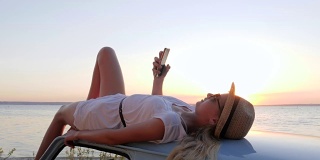 女孩手拿手机躺在车顶上，在手机上拍下女孩在夕阳下的照片