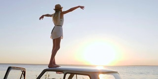 快乐的旅行，女孩站在车顶，双手张开，欣赏日落，自由的快乐在海边，年轻女子站在举起的手