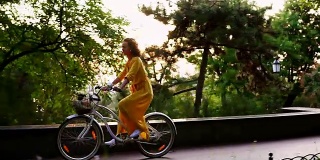 美丽微笑的女人穿着黄色长裙骑着一辆城市自行车，里面有一个篮子和鲜花在黎明。镜头光晕。替身。Slowmotion