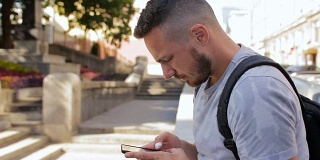 一个留着胡子的年轻人坐在外面的台阶上，用手机查看邮件