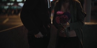 年轻夫妇在晚上站在城市的特写。男人送花给女人，在市中心见面