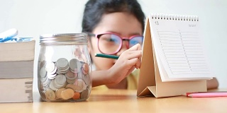 亚洲小女孩用桌子上的玻璃罐在日历上写字，隐喻省钱的概念