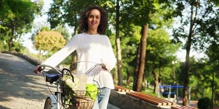微笑迷人的女人穿着白色的t恤和蓝色的牛仔裤走在她的城市自行车的车把和鲜花在篮子里在城市清晨。快乐的女人享受她的假期