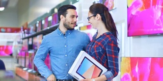 一对年轻夫妇在电子商店购物，他们已经为自己挑选了最新的平板电脑。商店是明亮的，现代的，有所有最新的电子设备。