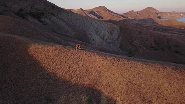 空中拍摄从飞行的无人机，一对年轻的游客正在享受美丽的海景日落慢动作，而乘坐的ATV运输。一对夫妇在海边的小山上骑着四轮自行车。4 k