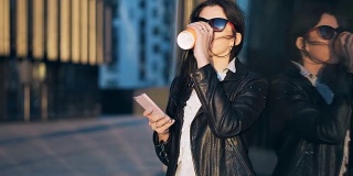 年轻迷人的女商人一边喝着咖啡，一边在有写字楼的城市里使用智能手机