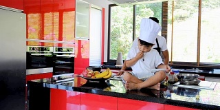 私人泰国厨师烹饪，他的小儿子在厨师帽旁边坐在桌子上的现代风格的家庭厨房。孩子在吃，爸爸在做饭
