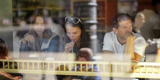 三位快乐的朋友在咖啡馆的餐桌上享用鸡尾酒，度过美好的时光。透过窗户看到街上餐馆里的两个女人和一个男人