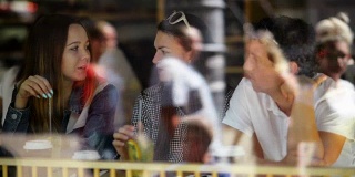 一个黑发女人和两个朋友坐在咖啡馆，喝着饮料聊天。透过餐厅的窗户看到三个朋友