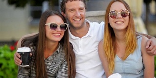 英俊的男人拥抱着两个戴着时尚太阳镜的漂亮女孩。三个微笑的朋友在室外看着相机摆姿势