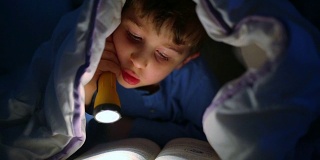 小男孩用手电筒在被子里看书