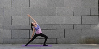 在灰色的城市背景下，一名女子在粉红色的垫子上练习瑜伽健身运动