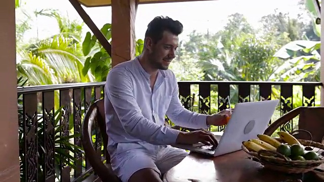 在热带森林的阳台上坐着吃早餐的休闲商人在用笔记本电脑工作，年轻的小伙子在打字