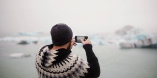 年轻时尚男子站在冰岛Jokulsalon冰礁湖的岸边，用智能手机拍照
