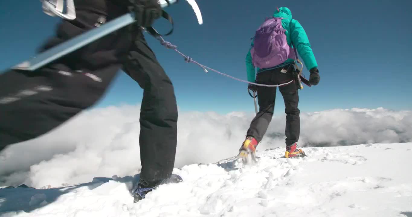 两名女性登山运动员在云层之上的高海拔处，在风吹过的冰川上穿越山脊。