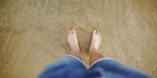 人的脚被海浪洗干净，埋在沙子里。一个穿着蓝色牛仔裤的白人男子把脚上的水刷掉，进入了大海。大海或海岸上的男性腿部特写。