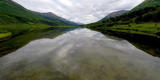 在阿拉斯加，一架4k无人机拍摄的视频显示，车辆和卡车在右侧风景优美的阿拉斯加高速公路上行驶时，从一片平静的玻璃湖向乡村山区行驶。阿拉斯加运输系统中的卡车和小汽车