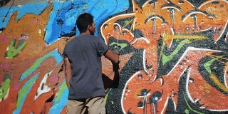 涂鸦艺术家用气溶胶作画。拿着喷雾器的人。缓慢的运动。年轻的城市画家在夏日的阳光下，在城市的街道墙上画上五颜六色的涂鸦。后视图，总体规划