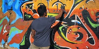 涂鸦艺术家用气溶胶作画。拿着喷雾器的人。缓慢的运动。年轻的城市画家在夏日的阳光下，在城市的街道墙上画上五颜六色的涂鸦。万向节。后视图，总体规划