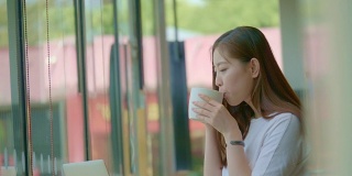 亚洲年轻女子在一个咖啡馆打电话，看着她的笔记本电脑屏幕在她的咖啡休息时间