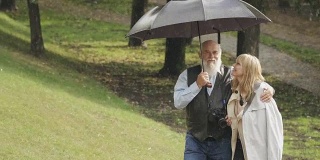 一位快乐的年轻女子和一位老人在雨中雨伞下在公园散步