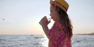 妈妈在户外给女儿喂水，小女孩在海边喝干净的水，可爱的孩子在背光下放杯子