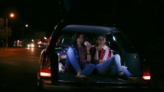 两个迷人的女朋友坐在敞开的后备箱，在城市的夜晚聊天和吃汉堡。Slowmotion拍摄视频素材模板下载