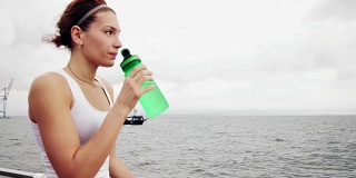 口渴的健身妇女休息休息，用水瓶饮用训练后。在海边训练的美女