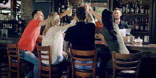 一群朋友在餐厅庆祝生日聚会，用鸡尾酒碰杯。坐在吧台的人，酒保坐在桌子的另一边，后面是箱子