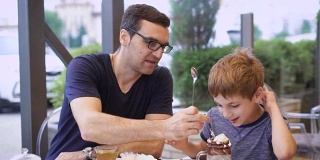 一位年轻的父亲尝试他儿子的牛奶鸡尾酒与冰淇淋和巧克力，儿子笑了，一家人正在餐厅用餐