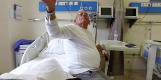老人躺在病床上，手上拿着点滴换电视频道。术后恢复