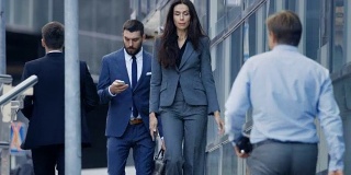 男和女商人走在繁忙的大城市街道上。他们都穿着时尚的灰/蓝西装，男人拿着包。他们急着去参加投资者会议。