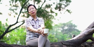 孤独的亚洲老年妇女坐在公园里独自喝咖啡，看起来很悲伤，思考着生活