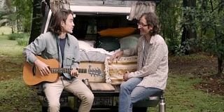 两个留着长发拿着吉他的男人坐在一辆卡车的后挡板上聊天
