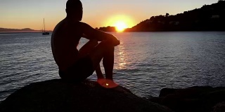 欣赏日落。一个男人坐在岩石沙滩上看风景