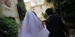一对新婚夫妇背对着陌生的新娘和新郎走在老城狭窄的街道上慢镜头。在意大利或欧洲的法国，穿着白色礼服和面纱的女人和穿着黑色礼服的男人手牵着手一起去教堂