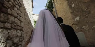 一对新婚的丈夫和妻子走在意大利古镇的一条认不出来的狭窄街道上结婚的慢镜头。年轻的家庭新娘与丈夫手挽面纱走向未来