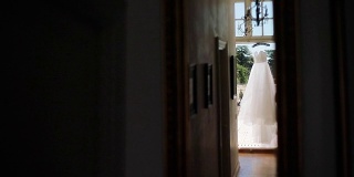 白色的衣服挂在阳台灯门口等着穿。从昏暗的走廊房间看到挂在衣架上的浅白色晚礼服或婚纱。新娘时尚设计清晨新娘夏日衣橱晾晒