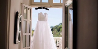 白色连衣裙挂在专门的衣架上挂在门口的露台上。由丝绸或雪纺织物制成的轻质夏季礼服在阳光明媚的早晨阳台等待新娘。婚礼时尚剪裁设计工作室