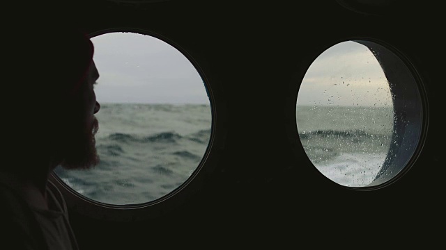 在波涛汹涌的海面上，一名长胡子的水手站在一艘船的舷窗前
