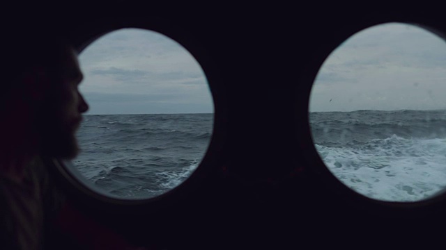 在波涛汹涌的海面上，一名长胡子的水手站在一艘船的舷窗前