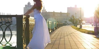 一个穿着白色衣服的美丽女孩在城市的河堤上迎接黎明。清晨，美好的一天。