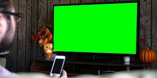 千禧一代男性一边在客厅看普通绿屏电视，一边用智能手机